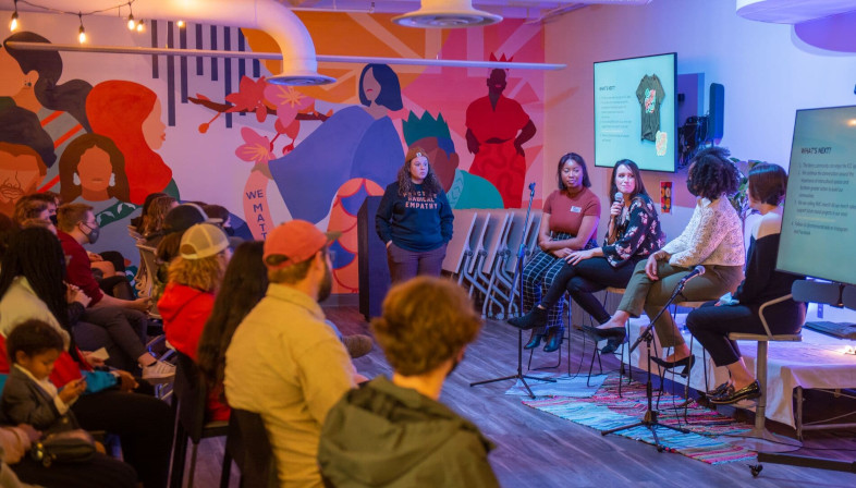 Four seated students present a new mural to a seated audience