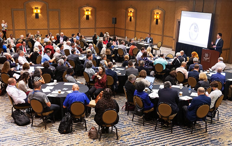 participants seated at round table looking at screen and presenter at podium situated at the front of the room