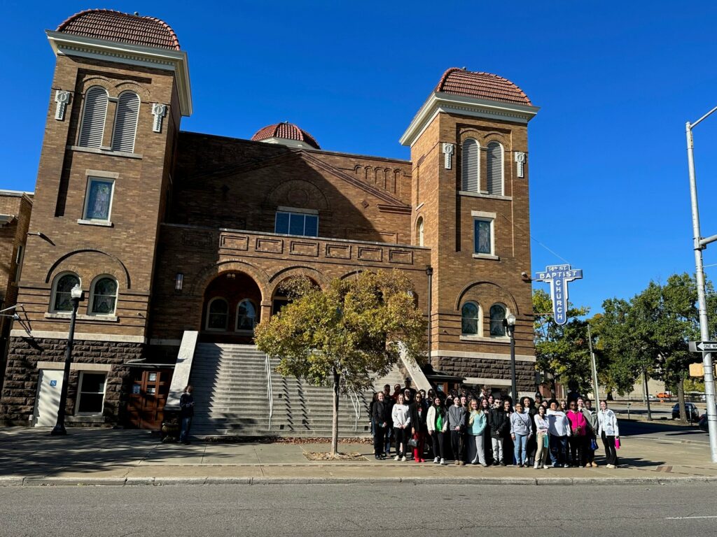 A group of students, faculty, and staff, stand in on the sidewalk front of a brown brick building- the 16th Street Baptist Church in Birmingham, Alabama.