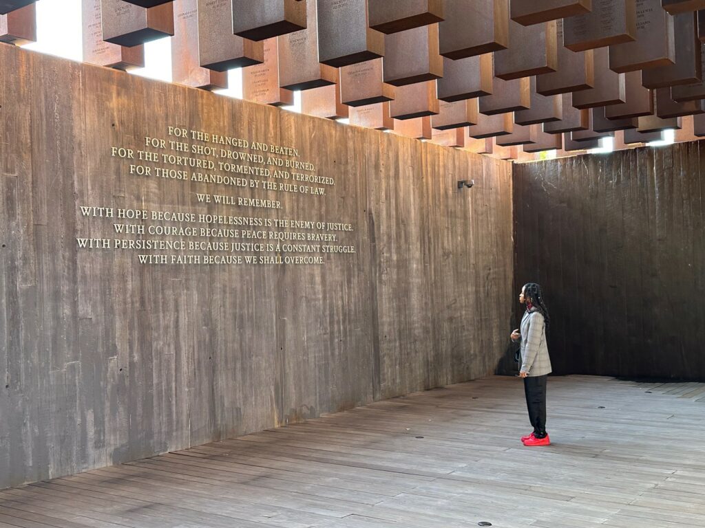 Student stands in the National Memorial for Peace and Justice in Montgomery, Alabama. two concrete walls, the concrete floor, and the hanging concrete sculptures overhead are in view. A student stands in the scene, reading text that is raised off the wall.