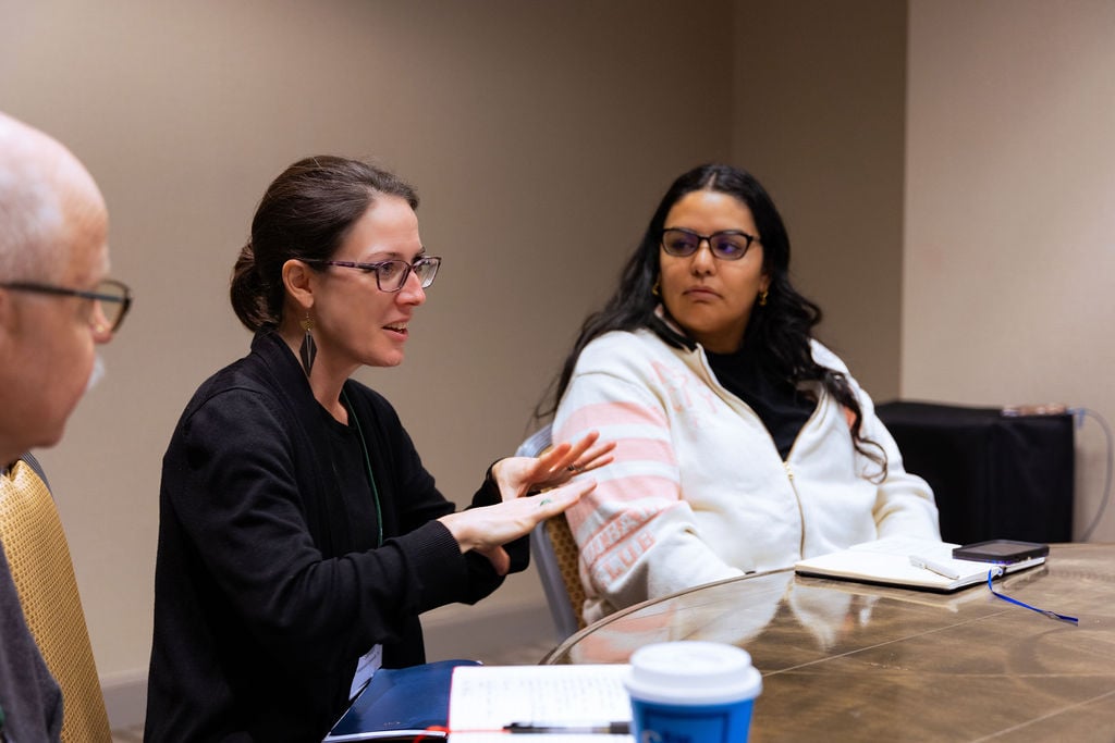 A woman seated at a table of gathering participants, talking. Other participants are listening attentively.