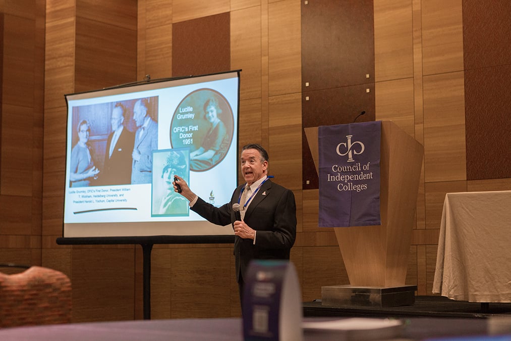 Speaker presents whiel standing in front of projection screen holding a slide advancer