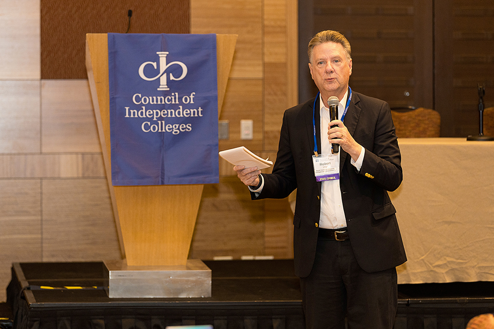 Speaker presents in front of podium while holding microphone and notepad