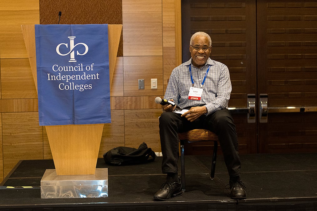 Speakers sits on stage next to podium smiling and holding a microphone