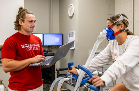 Student holding laptop while another student rides exercise bike wearing oxygen mask