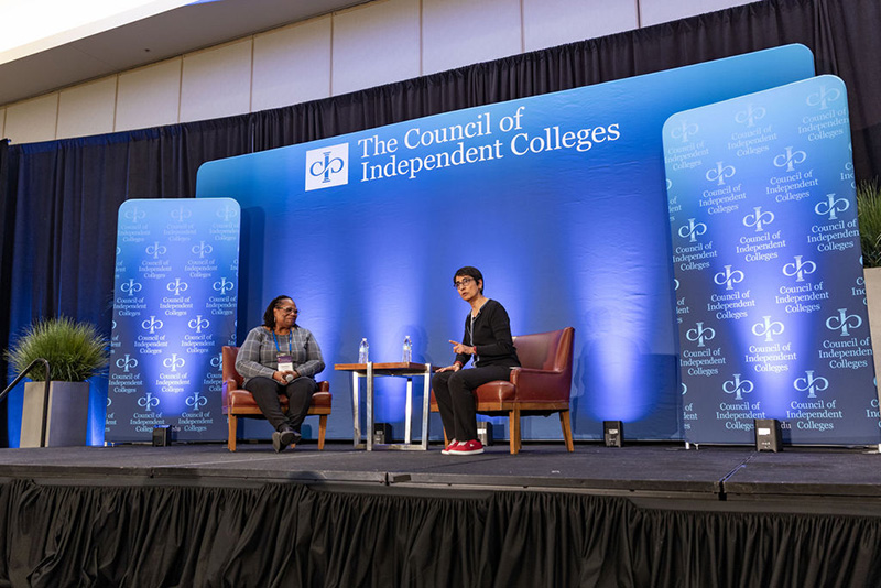 Two speakers present while seated on chairs on stage