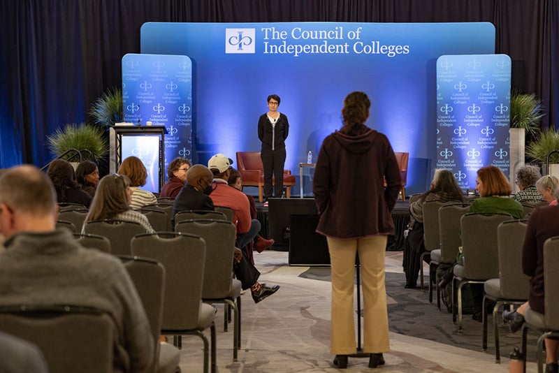 Irshad Manji stands on stage while participant asks a question at a standing microphone between rows of seated participants