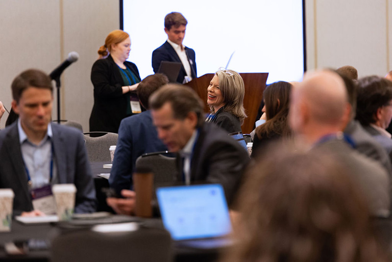 Two speakers stand at podium while participants discuss the interactive session topic at roundtables