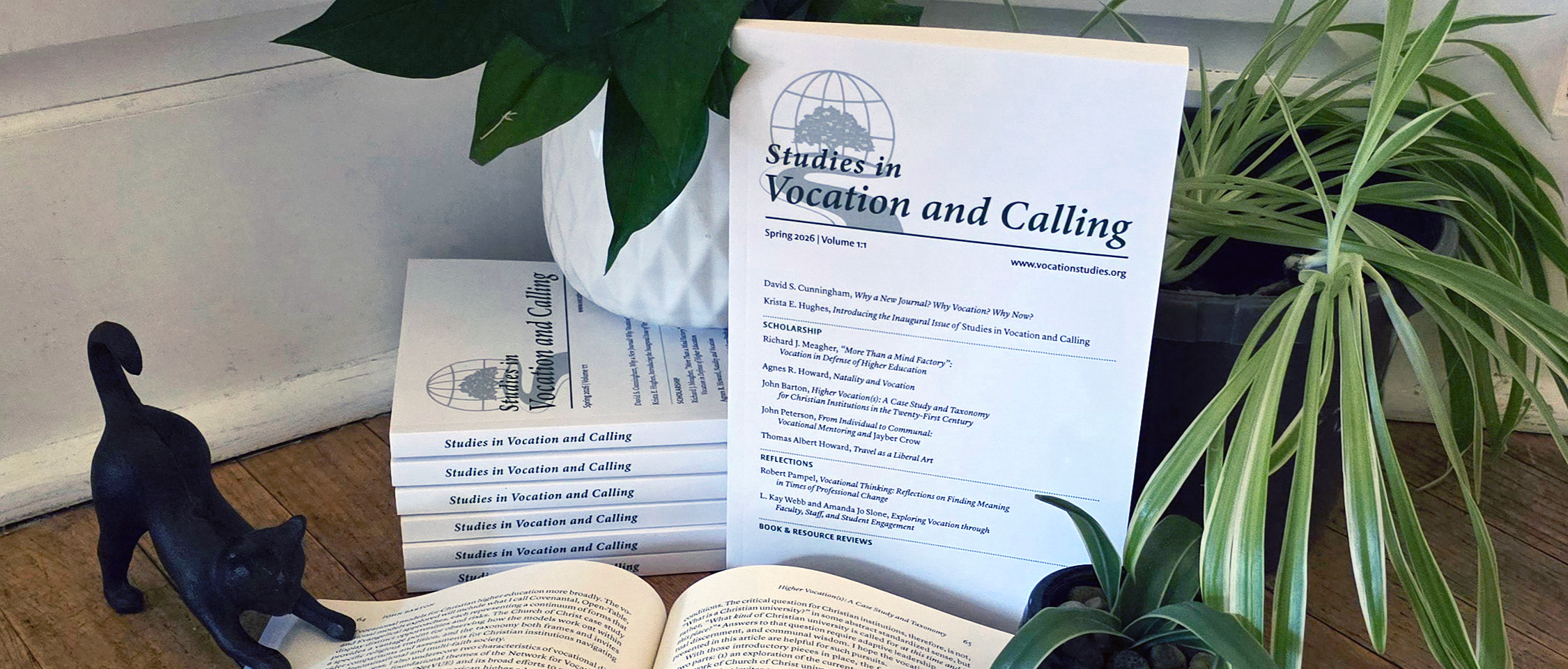 Copies of "Studies in Vocation and Calling" stacked with one placed upright and facing the viewer. The books are bordered by plants. One book is laid open in the foreground, held open by a stone cat desk ornament.